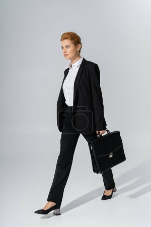 volle Länge der selbstbewussten Geschäftsfrau, die nach vorne schaut, während sie mit Aktentasche auf grauem Hintergrund geht