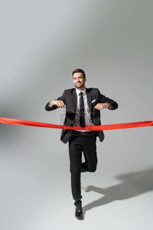 full length of happy businessman in black suit running and crossing red ribbon on grey background