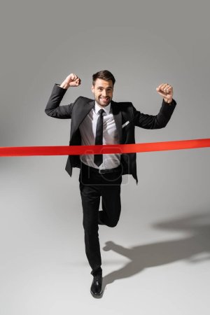 full length of overjoyed businessman crossing finish ribbon while running and showing win gesture on grey background