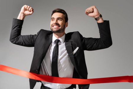 Photo for Young successful businessman crossing red finish ribbon and showing triumph gesture isolated on grey - Royalty Free Image