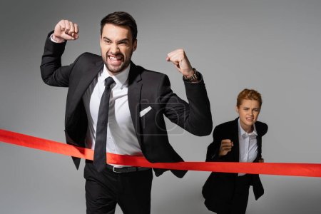 overjoyed businessman in black suit showing win gesture near sad woman running on grey background