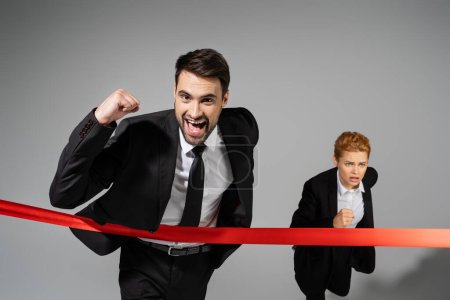 successful businessman looking at camera near finish ribbon and displeased businesswoman running on grey background