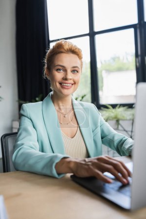 successful businesswoman in stylish blazer typing on laptop and smiling at camera in office