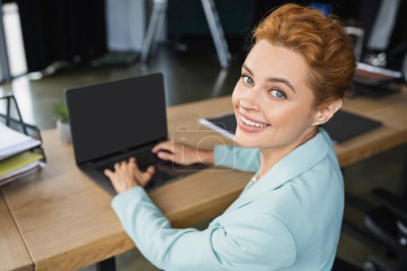 Téléchargez les photos : Joyeuse rousse femme d'affaires tapant sur ordinateur portable flou avec écran blanc et souriant à la caméra dans le bureau - en image libre de droit