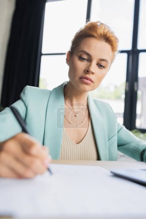 concentrated redhead businesswoman in stylish blazer writing in office on blurred foreground