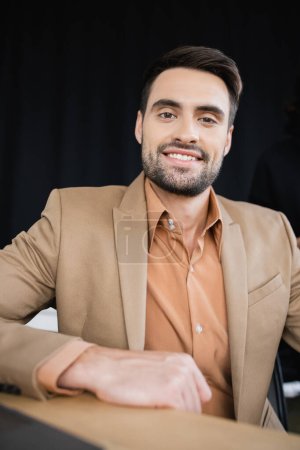 bearded businessman in beige blazer smiling at camera while sitting in office