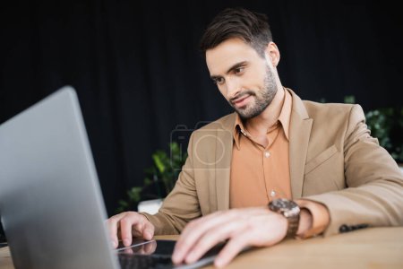 smiling businessman in beige blazer working on blurred laptop in office