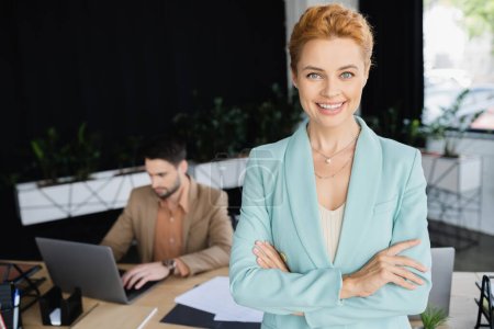 pleased businesswoman in trendy blazer looking at camera near colleague working on laptop in office