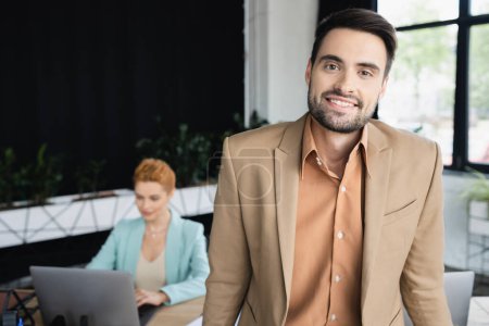 Photo for Bearded businessman in stylish blazer smiling at camera near blurred colleague working on laptop in office - Royalty Free Image