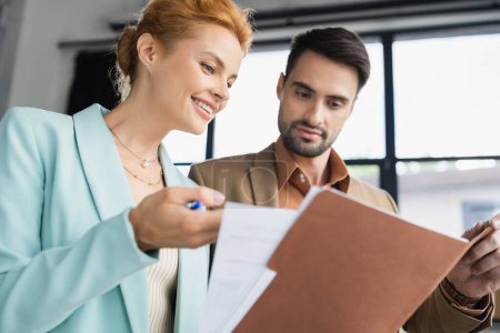 cheerful redhead businesswoman showing documents to bearded colleague in office