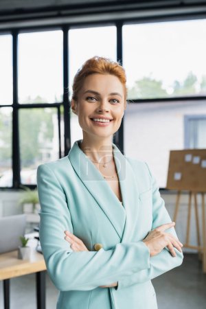 fröhliche rothaarige Geschäftsfrau in trendigen Klamotten blickt im Büro in die Kamera