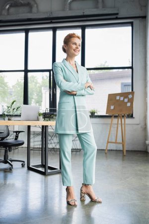 longitud completa de alegre pelirroja mujer de negocios en elegantes pantalones de pie con los brazos cruzados en la oficina moderna