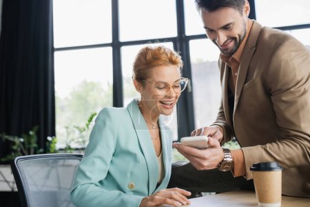 positive manager showing mobile phone to businesswoman in eyeglasses in office