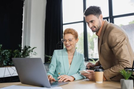 bearded businessman with coffee to go near manager in eyeglasses working on laptop in office