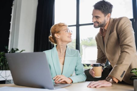mujer de negocios feliz en gafas de trabajo en el ordenador portátil y mirando colega barbudo con café para ir en la oficina