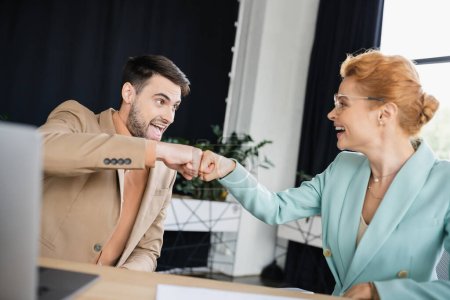 cheerful business partners giving high five near blurred laptop in office