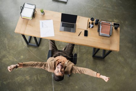 top view of overjoyed businessman showing win gesture near devices and documents on desk in office