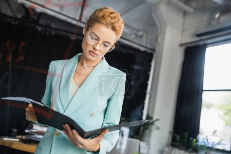 Nachdenkliche rothaarige Geschäftsfrau mit Brille und schickem Blazer blickt im Büro auf Ordner mit Dokumenten
