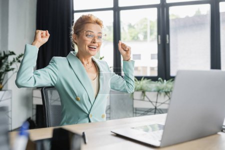 Téléchargez les photos : Joyeuse rousse femme d'affaires dans les lunettes montrant geste de victoire près d'un ordinateur portable au bureau - en image libre de droit