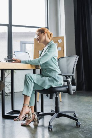 Téléchargez les photos : Pleine longueur de femme d'affaires en costume à la mode assis sur le lieu de travail dans le bureau - en image libre de droit