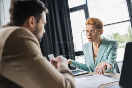Besorgte Geschäftsfrau mit Brille zeigt im Gespräch mit verschwommenem Amtskollegen auf Dokumente