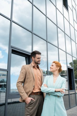 gente de negocios con estilo en blazers sonriendo el uno al otro cerca del edificio urbano con fachada de vidrio