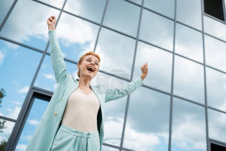 Tiefansicht einer überglücklichen Geschäftsfrau mit Siegergeste und Schreien in der Nähe eines Gebäudes mit Glasfassade