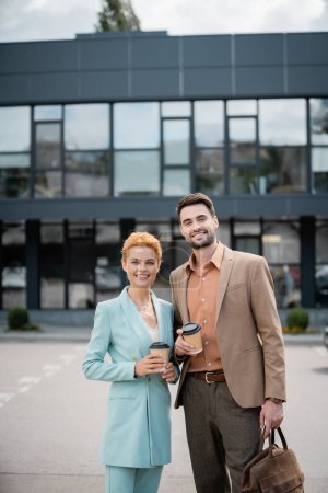 Photo for Cheerful and stylish business colleagues with takeaway drinks looking at camera near modern city building on blurred background - Royalty Free Image