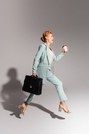 Foto de Longitud completa de mujer de negocios sorprendida en traje elegante levitando con café para llevar y maletín negro sobre fondo gris - Imagen libre de derechos