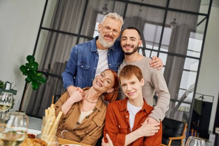 fröhlicher schwuler Mann mit Eltern und Freund, die sich umarmen und in die Kamera lächeln, in der Nähe des servierten Abendessens im Wohnzimmer
