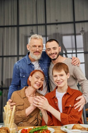 zufriedenes Paar mittleren Alters mit Sohn und seinem schwulen Partner, die sich umarmen und in die Kamera lächeln, in der Nähe köstlicher Mahlzeiten auf dem Tisch