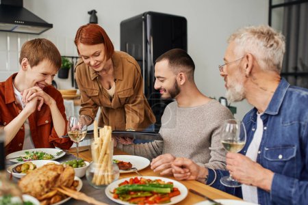 pelirroja mujer sosteniendo familia foto álbum cerca de hijo con gay pareja durante delicioso cena en cocina