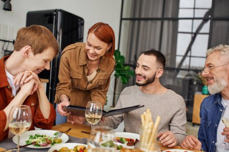 happy redhead woman showing family photo album to son with gay boyfriend during supper at home
