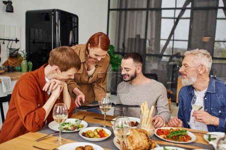 redhead woman showing family photo album to son with gay boyfriend near delicious supper at home