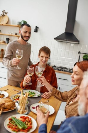 young gay couple with happy parents toasting with wine during family supper in kitchen