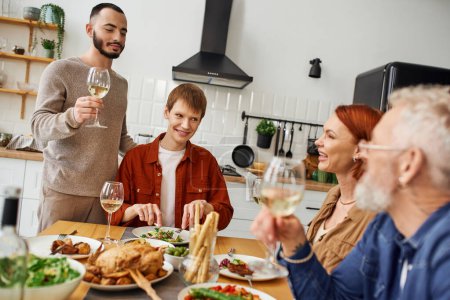 bärtiger schwuler Mann stößt mit Wein in der Nähe von Freund und glücklichen Eltern beim Familienessen in der Küche an