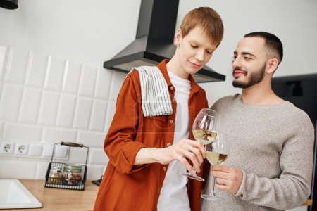 bärtiger schwuler Mann, der mit lächelndem Rotschopf in der Küche Weingläser klingelt