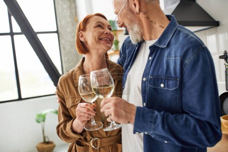 stylish and joyful couple clinking wine glasses and smiling at each other in kitchen
