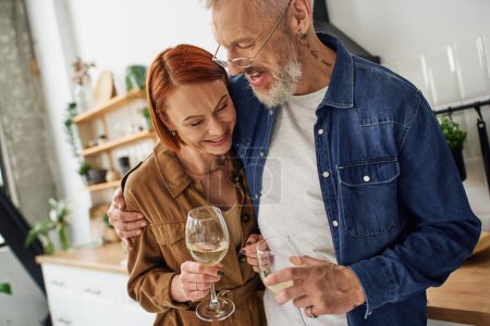 cheerful bearded man embracing redhead wife while holding wine glasses in kitchen