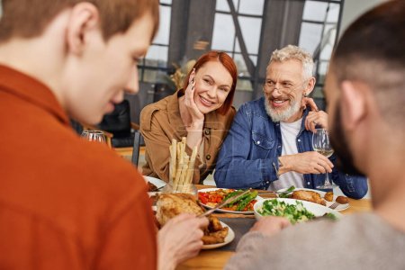 happy couple looking at son with gay partner having delicious supper in living room
