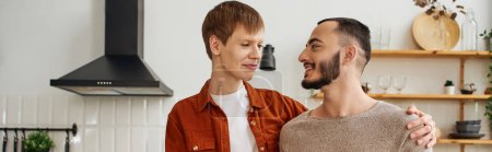 young and cheerful gay couple smiling at each other in kitchen, banner