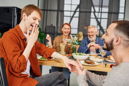 surprised man covering mouth while boyfriend making marriage proposal near happy parents during family supper in living room
