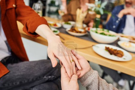 abgeschnittene Ansicht von homosexuellen Mann hält Hand auf Freund im Ehering in der Nähe verschwommenes Abendessen und Eltern zu Hause