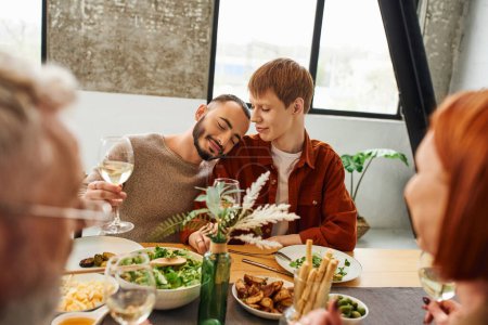 complacido hombre gay con copa de vino apoyado en pelirroja novio cerca borrosa padres y deliciosa cena en cocina