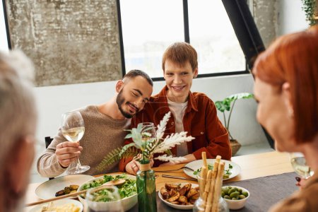 joyful bearded man with glass of wine leaning on happy gay partner during family dinner in kitchen