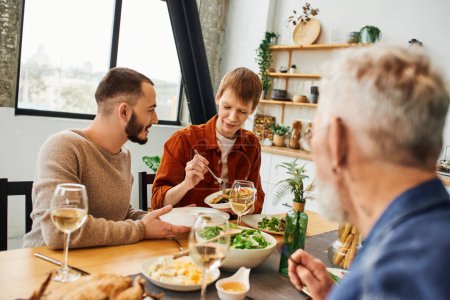 Téléchargez les photos : Jeune rousse homme servant un délicieux repas près partenaire gay pendant le dîner en famille dans la cuisine - en image libre de droit