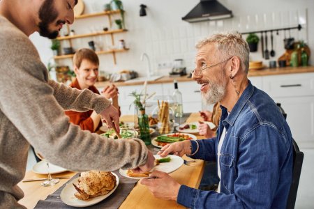 smiling bearded man looking at gay man serving chicken during supper with family in kitchen