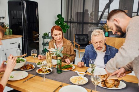 bearded gay man cutting grilled chicken during family dinner with boyfriend and parents in kitchen