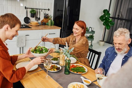 happy redhead woman serving salad during delicious supper with husband and son in modern kitchen