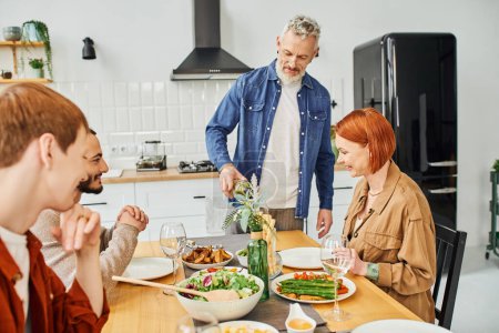 Photo pour Homme barbu verser du vin près de la famille heureuse ayant délicieux souper dans la cuisine moderne - image libre de droit